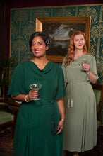 Load image into Gallery viewer, Two women standing indoors in elegant dresses, holding cocktail glasses. The woman in the foreground wears a dark green cowl-neck dress, and the woman behind her wears a light sage green midi dress.