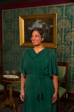 Load image into Gallery viewer, A woman wearing a dark green draped cowl-neck dress with a tie waist and tassel details, standing in a vintage room with patterned wallpaper and a framed painting.