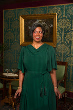 Load image into Gallery viewer, A woman with short gray hair wearing a dark green draped dress with a tie waist, standing in a vintage room with patterned wallpaper and a framed painting.