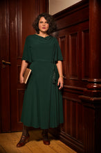 Load image into Gallery viewer, A woman stands in a wood-paneled room wearing a dark green midi dress with short sleeves, a draped neckline, and a self-tie waist belt. She holds a brown book and wears brown lace-up ankle boots.