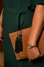 Load image into Gallery viewer, A close-up lifestyle shot of a person wearing a dark forest green dress and holding a brown leather clutch bag with black tassels.