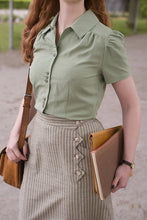 Load image into Gallery viewer, Person wearing a green shirt and gray skirt holding a brown bag and book outdoors.