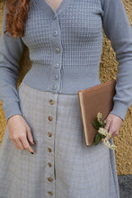 Load image into Gallery viewer, Person wearing a gray textured cardigan and skirt holding a brown folder with flowers against a textured yellow wall.