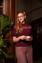 Load image into Gallery viewer, A woman with red hair and glasses wearing a burgundy short-sleeve knit sweater with a geometric pattern and mauve trousers, holding a teacup and saucer in a dimly lit indoor setting.