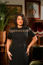 Load image into Gallery viewer, A woman with curly dark hair wearing a black short-sleeve dress with a geometric diamond pattern across the chest and a matching belt, standing in a vintage-style room with wood paneling, framed art, a potted plant, and a red velvet lamp.