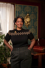 Load image into Gallery viewer, A woman stands indoors wearing a black short-sleeve knit top with a geometric diamond pattern in beige along the shoulders and chest, paired with dark gray plaid trousers. She is posed in a room with patterned teal wallpaper, a gold wall clock, a window with white curtains, and a wooden table with books.