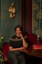 Load image into Gallery viewer, A woman sits at a wooden table in an ornate room with patterned wallpaper, a gold wall clock, and a red upholstered bench. She is wearing a black short-sleeve top with gold geometric embroidery on the shoulders and chest, paired with dark gray plaid trousers.