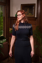 Load image into Gallery viewer, A woman with red hair and glasses wearing a dark navy blue short-sleeve dress with a white geometric diamond pattern across the chest and a matching belt, standing in a vintage-style room with wooden furniture and plants.