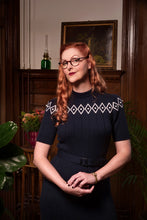Load image into Gallery viewer, A woman with red hair and glasses wearing a dark navy blue ribbed dress with a white geometric diamond pattern across the chest, standing in a vintage-style room with dark wood paneling.
