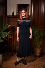 Load image into Gallery viewer, A woman with red hair and glasses wearing a dark navy blue midi dress with white geometric embroidery on the yoke, standing in a vintage room with wooden paneling and plants.