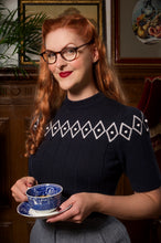 Load image into Gallery viewer, A woman with red hair and glasses wearing a navy blue short-sleeve knit sweater with a white geometric diamond pattern across the chest, holding a blue and white patterned teacup and saucer in a vintage-style indoor setting.