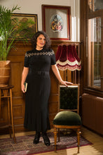 Load image into Gallery viewer, A woman stands in a vintage-style room wearing a black short-sleeve midi dress with a geometric pattern across the chest and a matching belt. She is posed next to a green velvet upholstered chair in a room with wooden paneling, framed art, and a patterned rug.