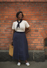Load image into Gallery viewer, Woman standing against a brick wall wearing a white top, navy skirt, and carrying a woven bag.