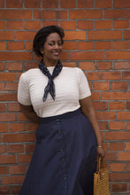Load image into Gallery viewer, Woman wearing a white top and navy skirt with a patterned scarf, standing against a brick wall.