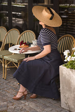 Load image into Gallery viewer, Woman sitting at an outdoor cafe table wearing a straw hat and dark dress.