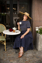 Load image into Gallery viewer, Woman in a striped shirt and navy skirt sitting at an outdoor cafe table, holding a cup.