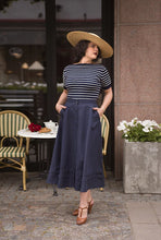 Load image into Gallery viewer, Woman in a striped shirt and navy skirt standing outdoors near a cafe.