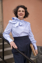 Load image into Gallery viewer, Woman wearing a blue striped blouse with a large bow and dark pants, standing against a beige wall.