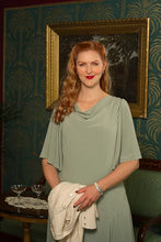 Load image into Gallery viewer, A woman with red hair stands indoors wearing a sage green draped dress and holding a cream-colored cardigan. She is posed in front of a patterned teal wall with a large gold-framed painting.