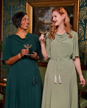 Load image into Gallery viewer, Two women standing indoors wearing elegant draped maxi dresses, one in deep teal and one in sage green, each holding a cocktail glass. The background features ornate green and gold patterned wallpaper and a large framed landscape painting.