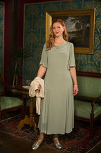 Load image into Gallery viewer, A woman stands in a vintage-style room wearing a sage green cowl-neck midi dress with short flutter sleeves. She holds a cream-colored jacket in one hand and wears silver heels, set against a background of patterned wallpaper, a green sofa, and a framed painting.