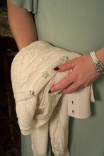 Load image into Gallery viewer, A close-up of a person holding a cream cable-knit sweater with star-shaped embellishments. The person is wearing a sage green dress and a pearl bracelet.