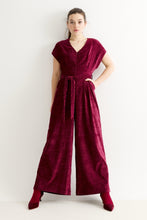 Load image into Gallery viewer, A woman modeling a deep burgundy velvet jumpsuit with a V-neck, short sleeves, a matching waist tie, and wide legs, paired with matching pointed-toe heels against a plain white studio background.