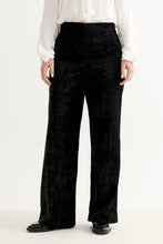 Load image into Gallery viewer, A person wearing black wide-leg trousers with a textured, velour-like fabric, styled with a white long-sleeve blouse and black shoes against a plain white studio background.