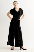 Load image into Gallery viewer, A woman modeling a black velvet short-sleeve jumpsuit with a V-neck, waist tie, and wide legs, paired with dark red heels, against a plain white studio background.