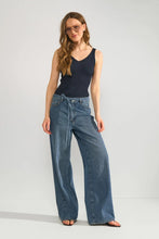 Load image into Gallery viewer, A woman modeling a pair of wide-leg blue denim jeans with a drawstring waist, paired with a dark navy ribbed tank top and flat sandals, against a light gray studio background.