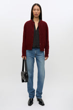 Load image into Gallery viewer, A model stands against a plain white studio background wearing a dark red knit cardigan over a black and white plaid shirt, blue jeans, and black shoes, while holding a small black handbag.
