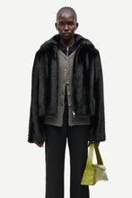 Load image into Gallery viewer, A model stands against a light gray studio background wearing a black faux-fur jacket over a gray knit cardigan and black pants, while holding a bright yellow-green handbag.