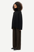 Load image into Gallery viewer, A model with voluminous dark curly hair is shown in profile wearing a black oversized turtleneck sweater and dark brown wide-leg trousers against a light gray studio background.