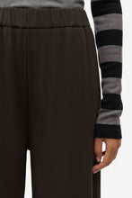 Load image into Gallery viewer, Close-up of a person wearing dark brown wide-leg pants and a gray and black striped long-sleeve top, with the hand holding the hem.