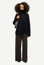 Load image into Gallery viewer, A model with an afro hairstyle stands against a light gray background wearing a black oversized turtleneck sweater, dark brown wide-leg trousers, and black shoes, while carrying a striped shoulder bag.