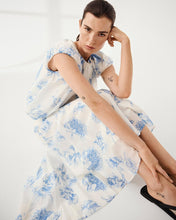 Load image into Gallery viewer, Woman wearing a white dress with blue floral pattern sitting on a white background